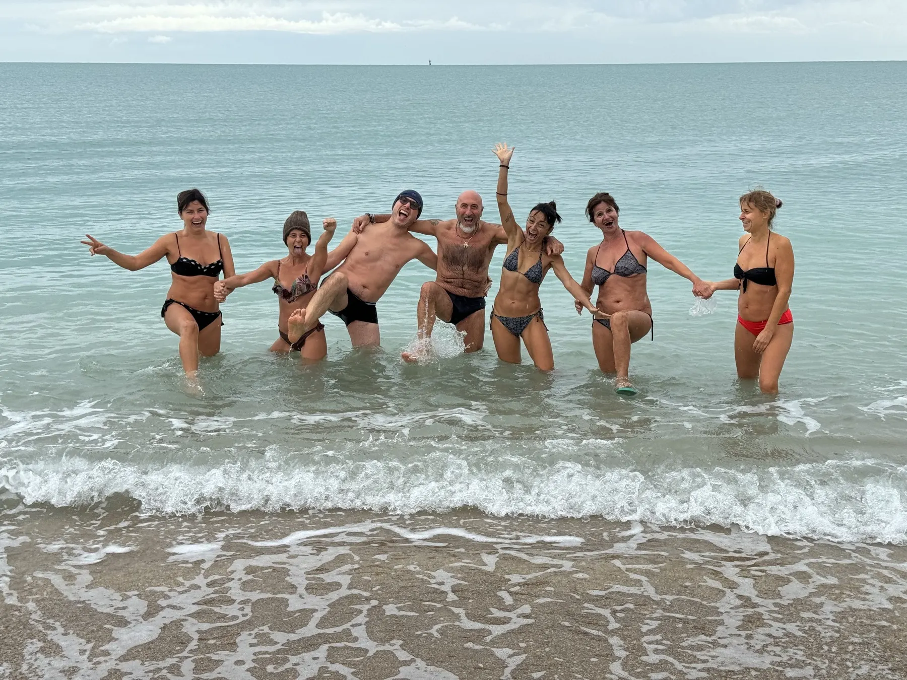 Un gruppo di persone in costume ride e salta tra le onde basse del mare in una giornata grigia